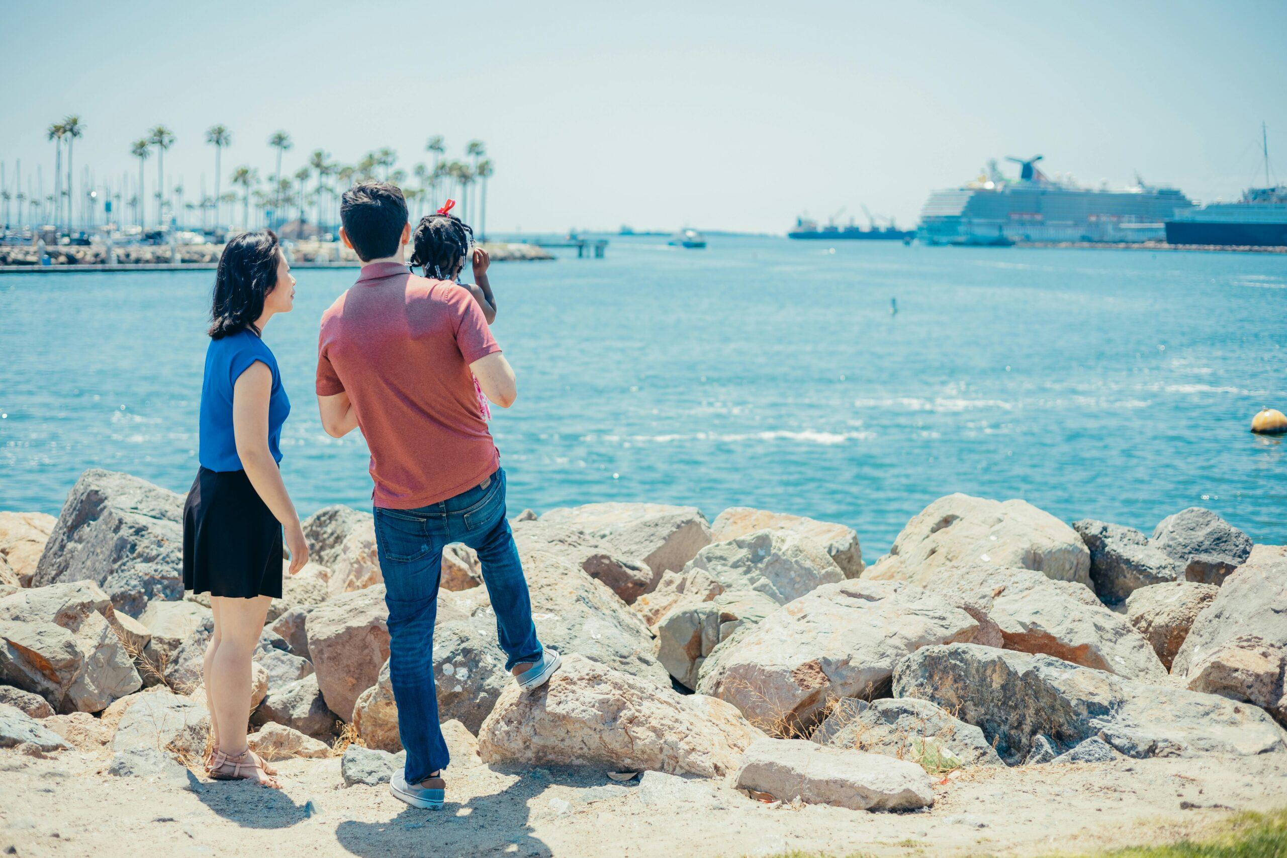 A multicultural family enjoys a sunny day by the sea with cruise ships in the background.